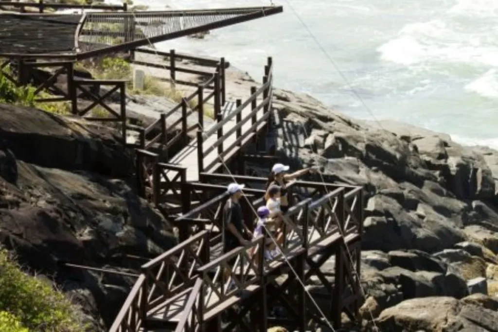 Passarelas de madeira sobre rochas no Museu Arqueológico ao Ar Livre Costão do Santinho, em Florianópolis, com pessoas explorando o local.
