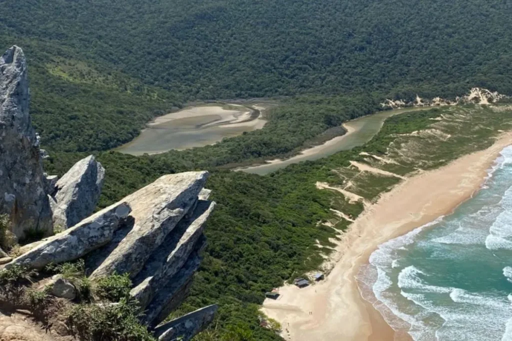 Vista do alto da Trilha do Morro da Coroa em Florianópolis, com formações rochosas, vegetação densa e praia ao fundo.